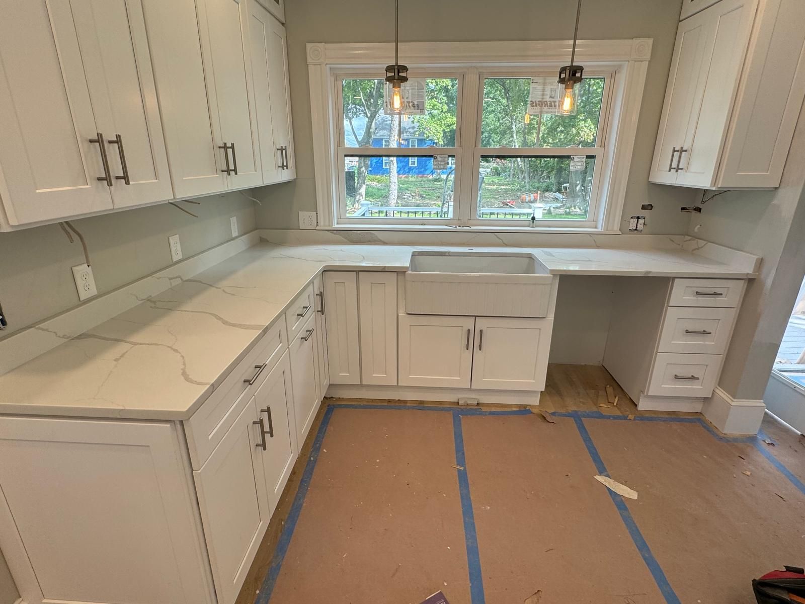 A newly renovated kitchen with white cabinets, light countertops, a farmhouse sink, and a large window.