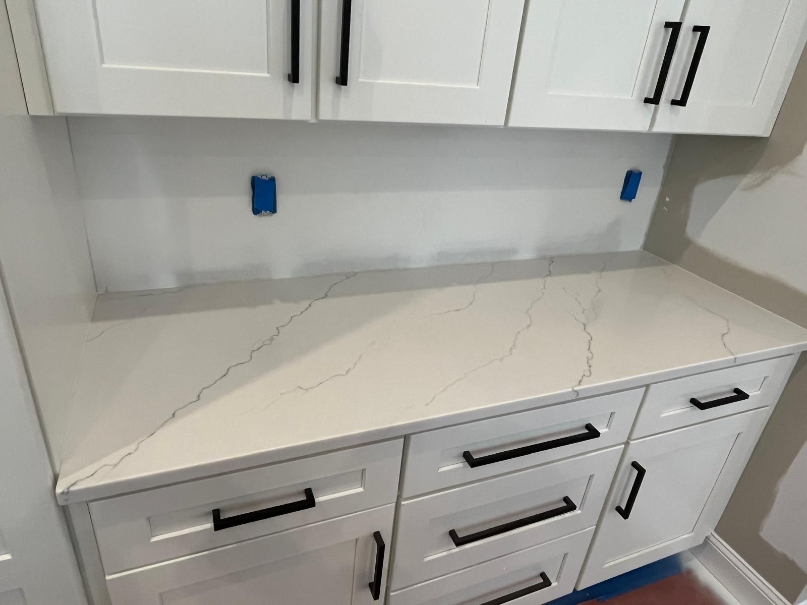 White cabinets with black handles and a light-colored countertop. Blue electrical outlets on the wall.