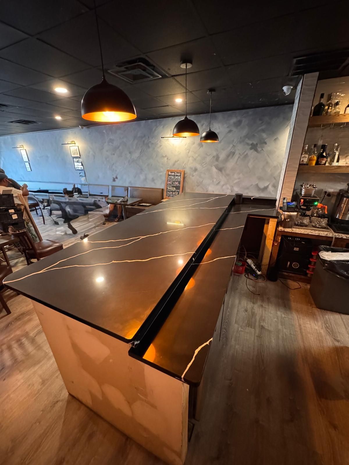 Bar with dark countertop, pendant lights, and unfinished wall; shelving with bottles to the right.