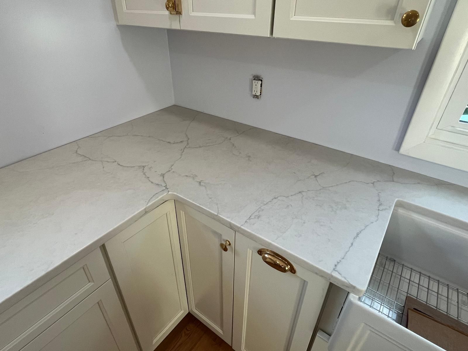 White kitchen cabinets and countertops with veining, near a window.