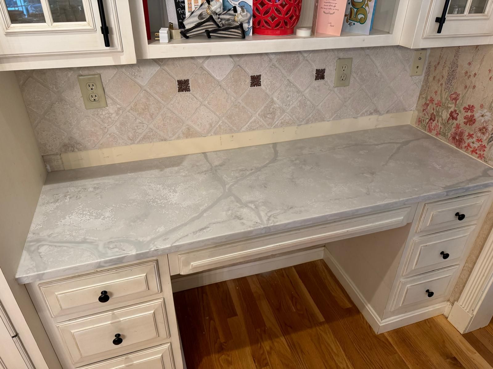 Built-in white desk with gray marble-look countertop and drawers, below cabinets with backsplash.
