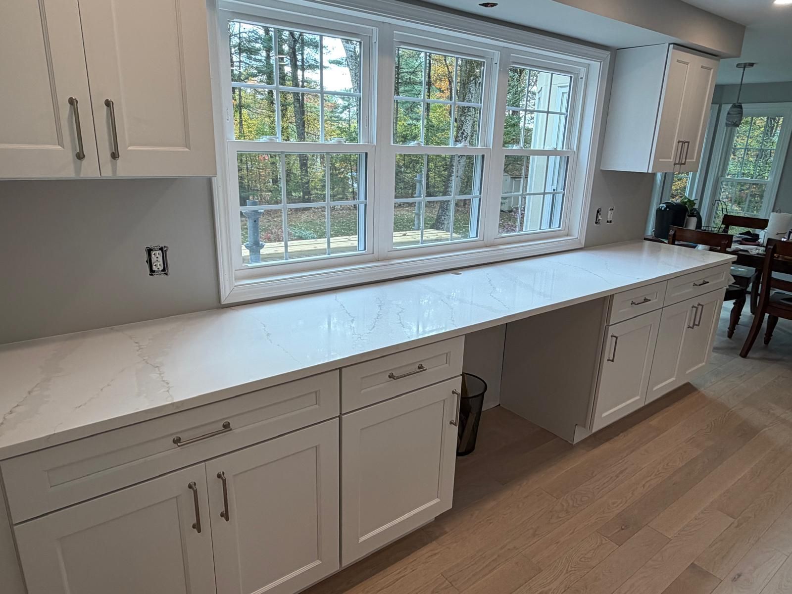 White kitchen cabinets with white countertops beneath a large window, light wood floors.