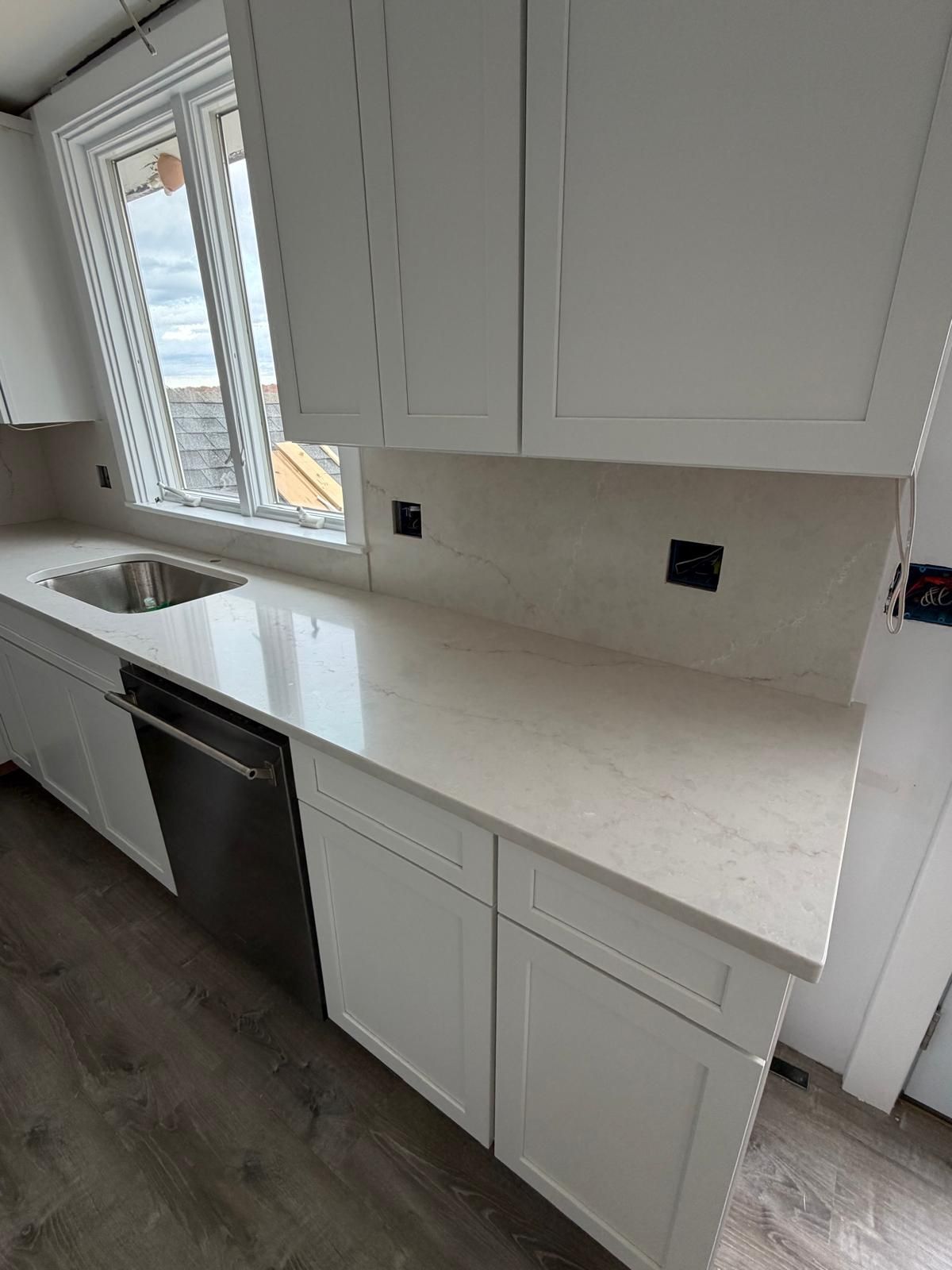 White kitchen cabinets and countertops with a stainless steel dishwasher and sink near a window.