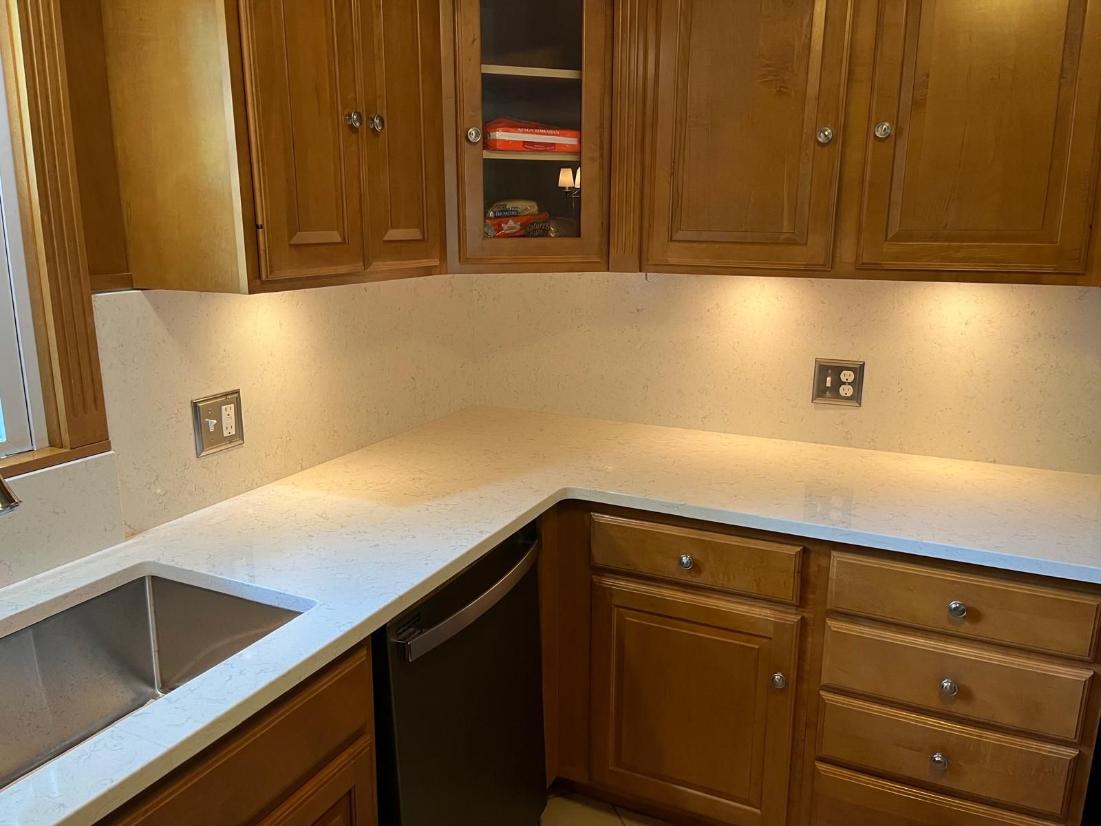 Kitchen with light wood cabinets, white countertops, and under-cabinet lighting. Stainless steel sink and appliances.
