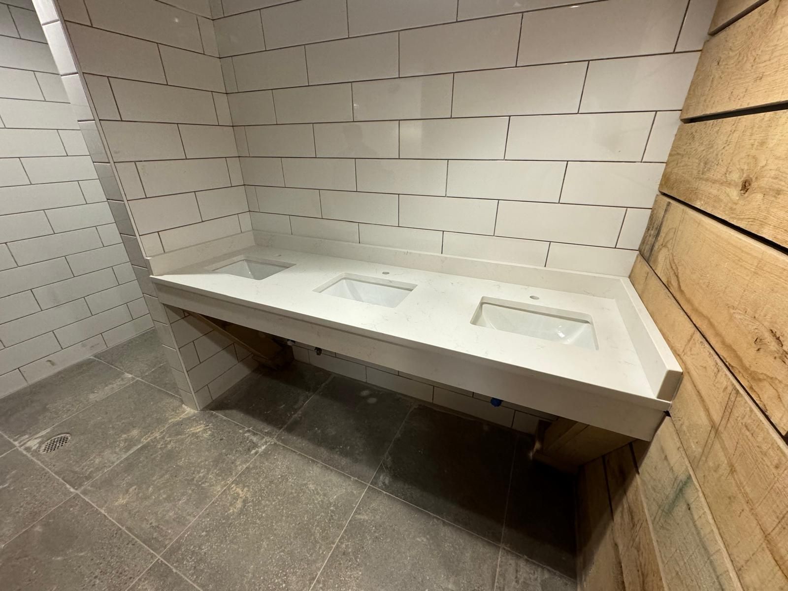 Bathroom with a white countertop and three sinks, white subway tile wall, and wood paneling on the right.