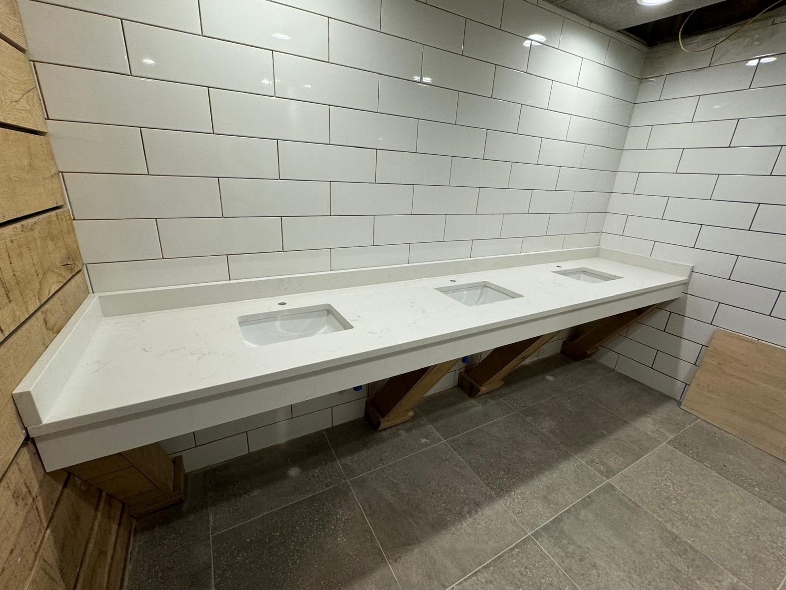 Long white countertop with three sinks and wooden supports, set against white brick tile wall.
