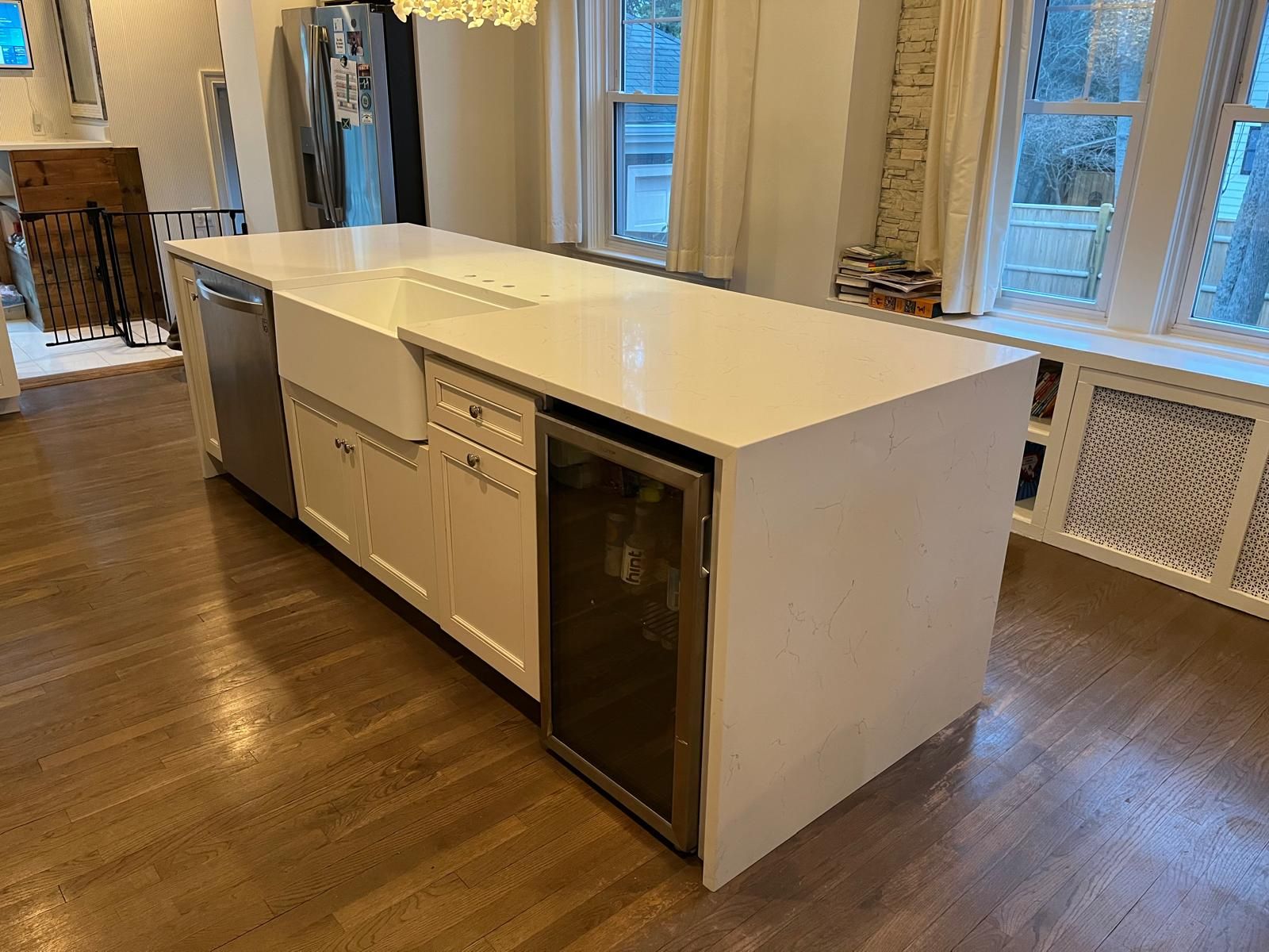 White kitchen island with a built-in sink, dishwasher, and wine fridge. Hardwood floor and windows.