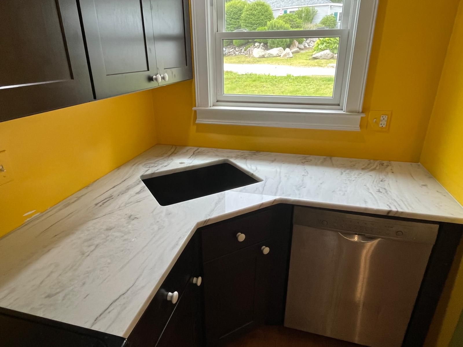 Kitchen counter with a sink and dishwasher. White marble-like countertop, dark cabinets, yellow walls.