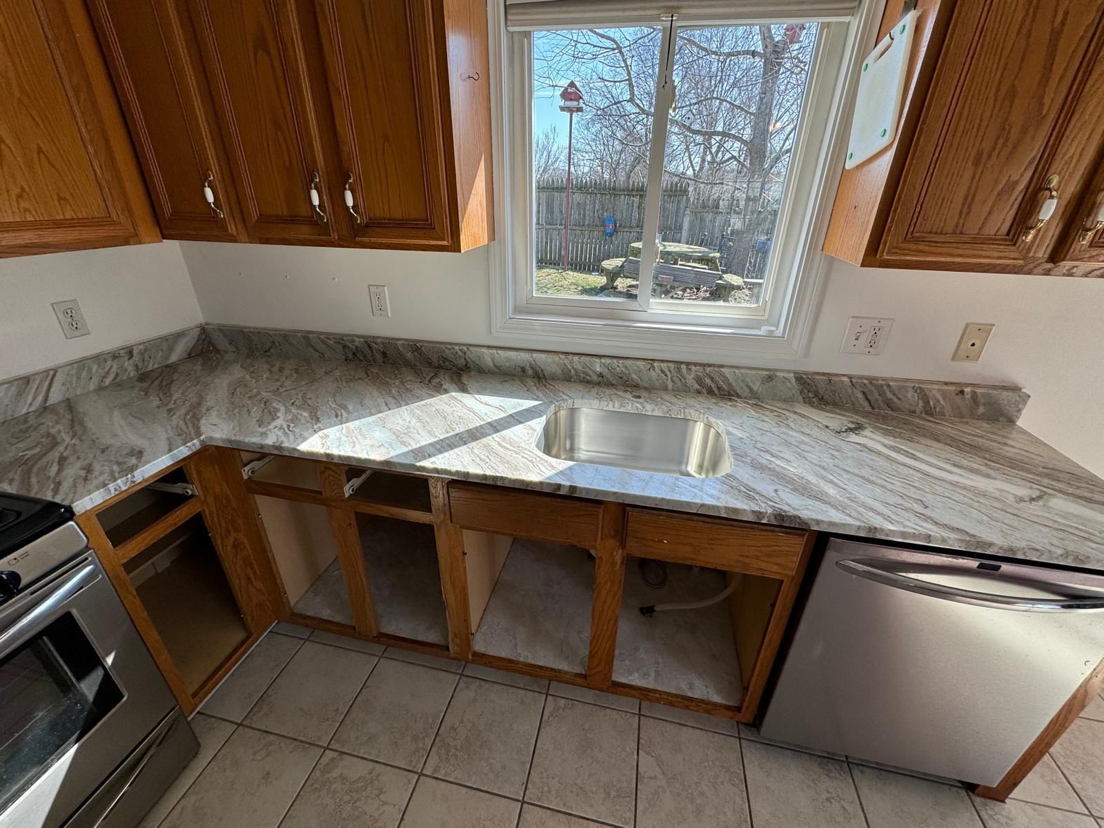 Kitchen with new granite countertops, cabinets, sink, and dishwasher under window; countertops are light gray.