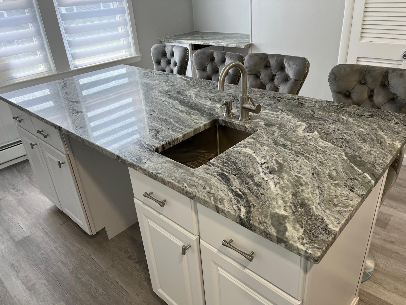 Kitchen island with granite countertop, sink, and white cabinets. Gray tufted chairs at a mirrored table.