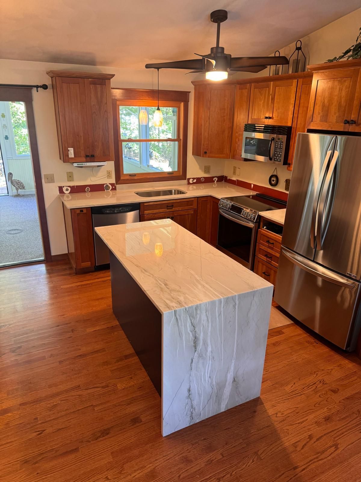 Kitchen with brown cabinets, stainless steel appliances, and a marble-topped island on a hardwood floor.