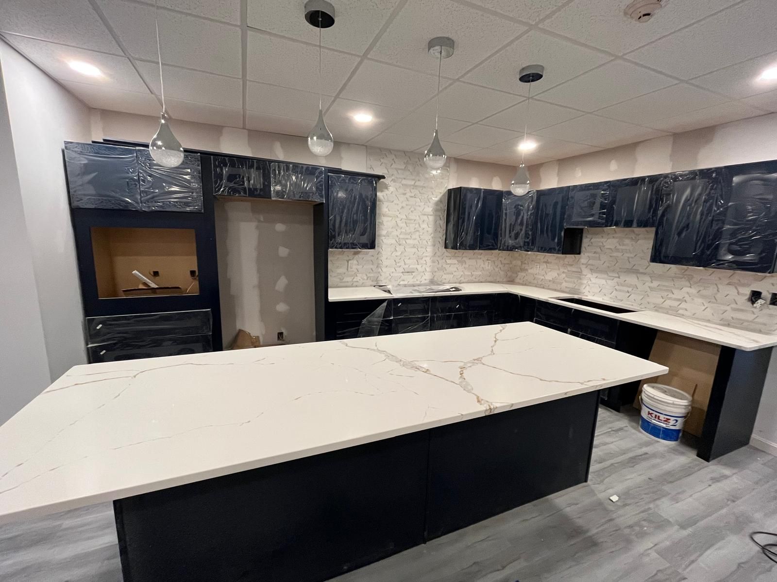 A kitchen with dark cabinets, a white countertop island, and light fixtures. Partially finished with protective plastic.
