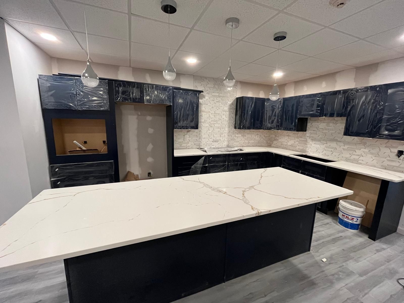Kitchen under renovation with dark blue cabinets, white countertop island, pendant lights, and light tile backsplash.