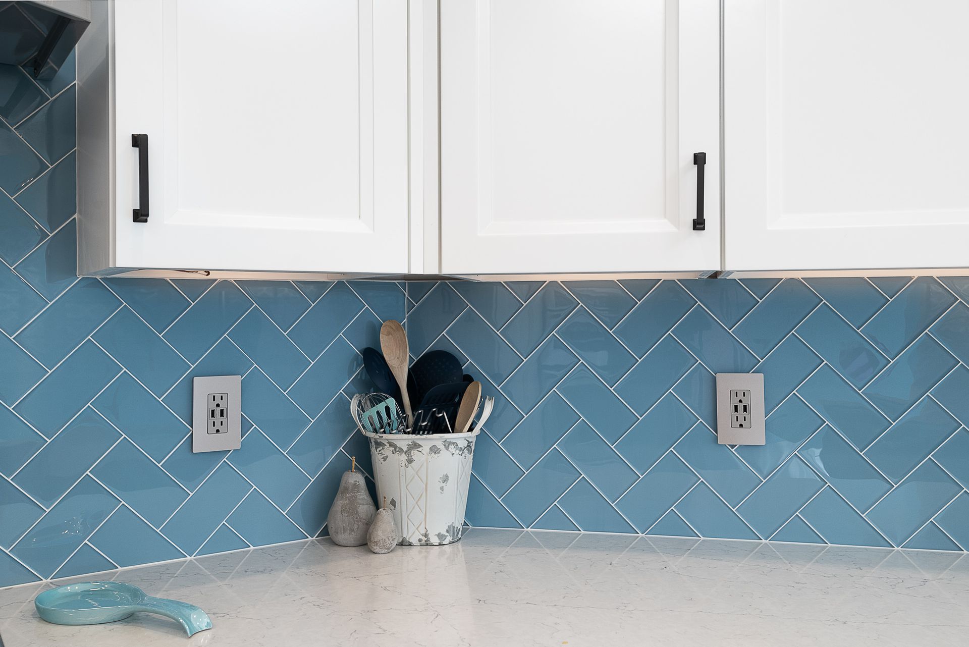 A kitchen with blue tiles and white cabinets and a bucket of utensils on the counter.