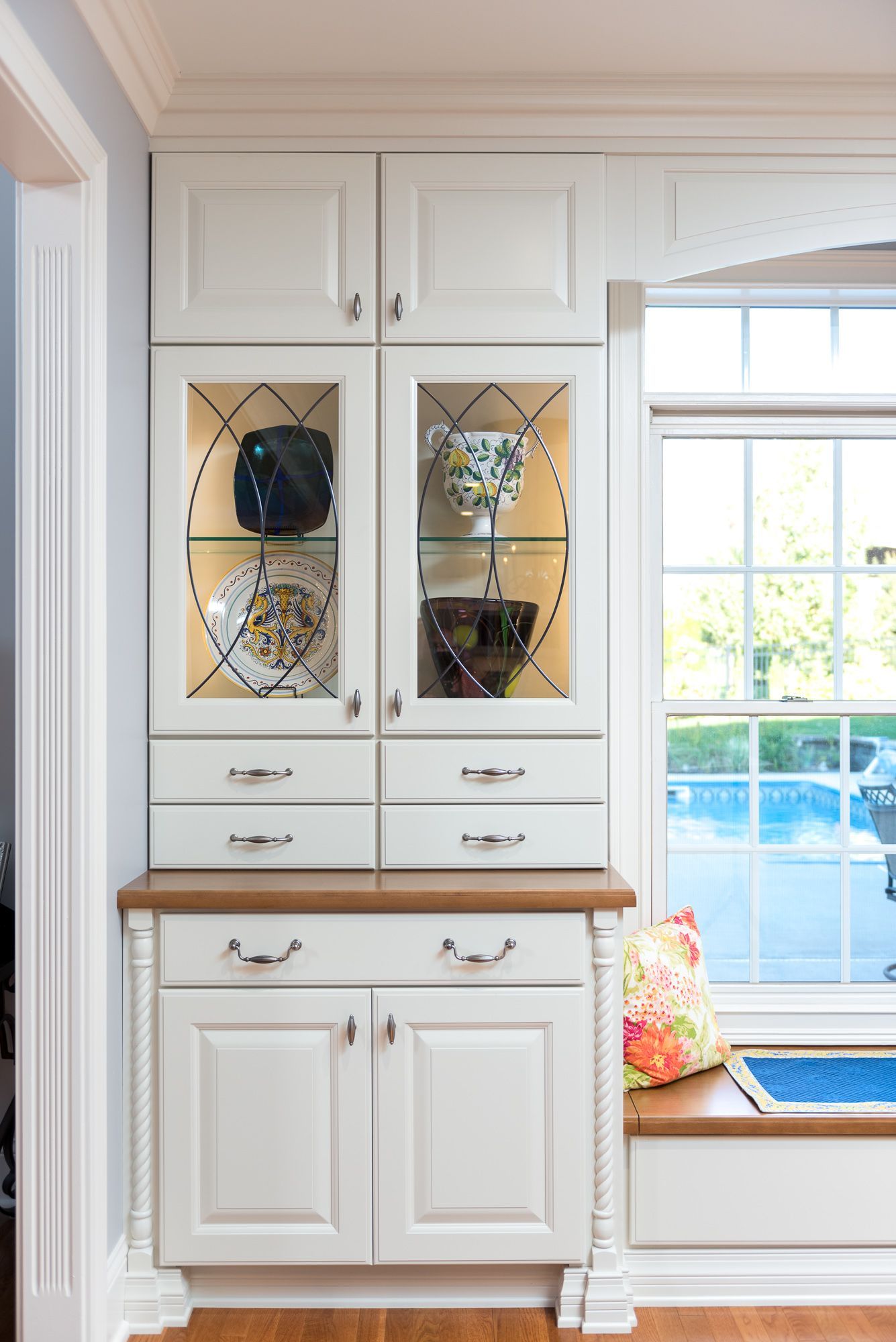 A white cabinet with a glass door and drawers in a kitchen next to a window.