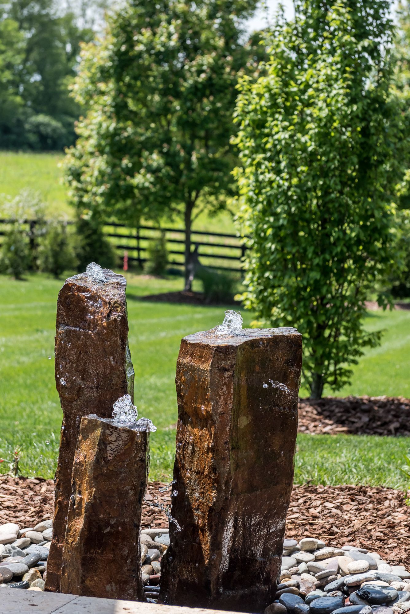 A water fountain made of rocks in a backyard with trees in the background.