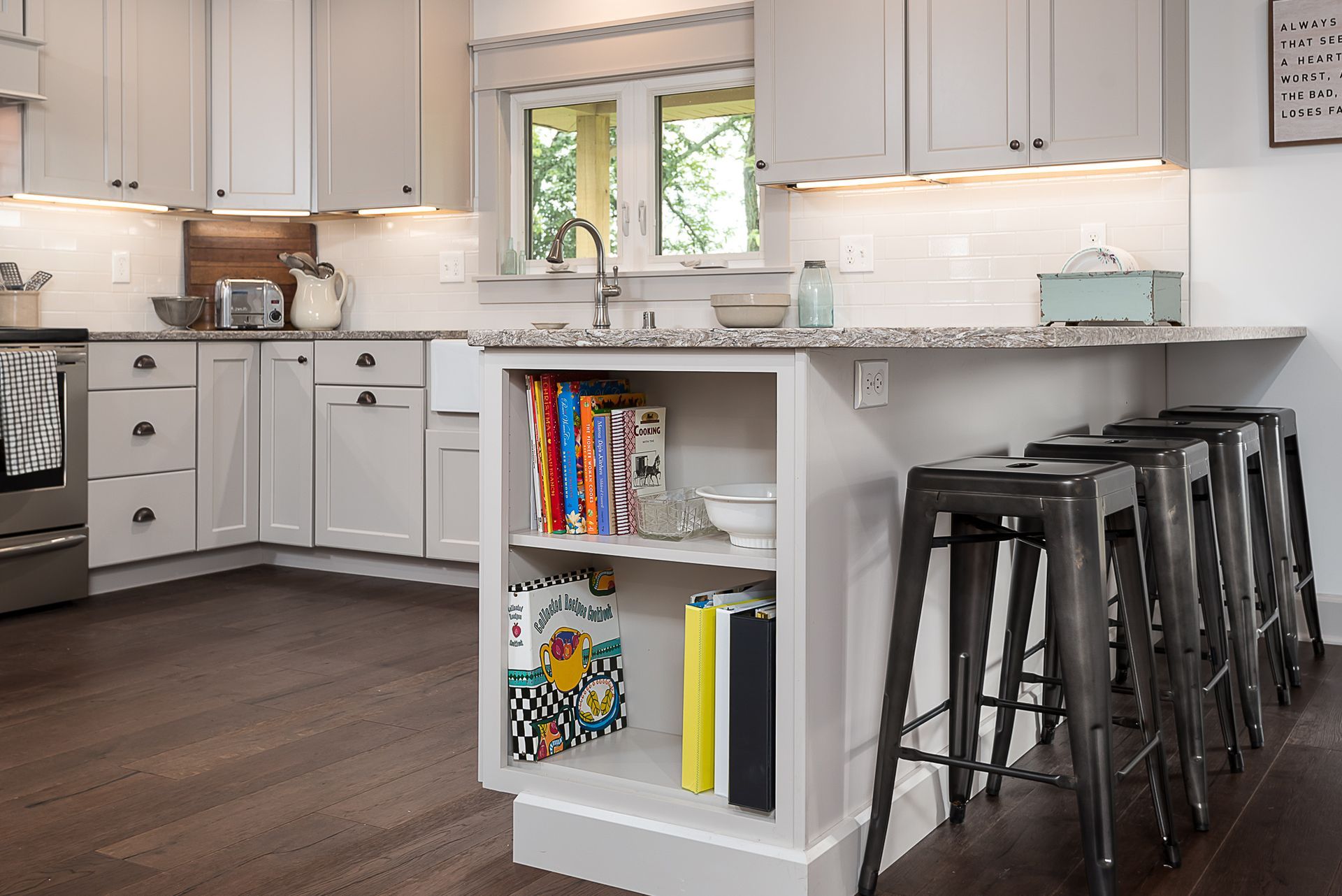 A kitchen with white cabinets and stools and a shelf with books on it.