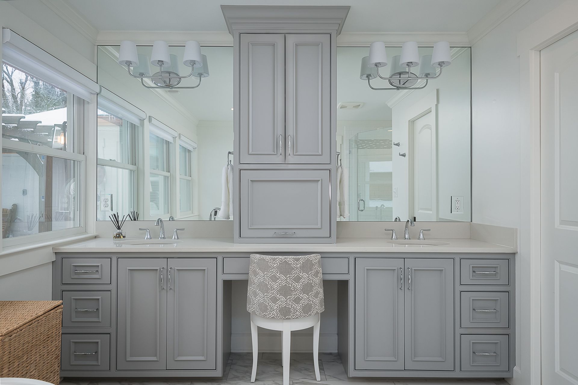 A bathroom with gray cabinets , two sinks , two mirrors and a chair.