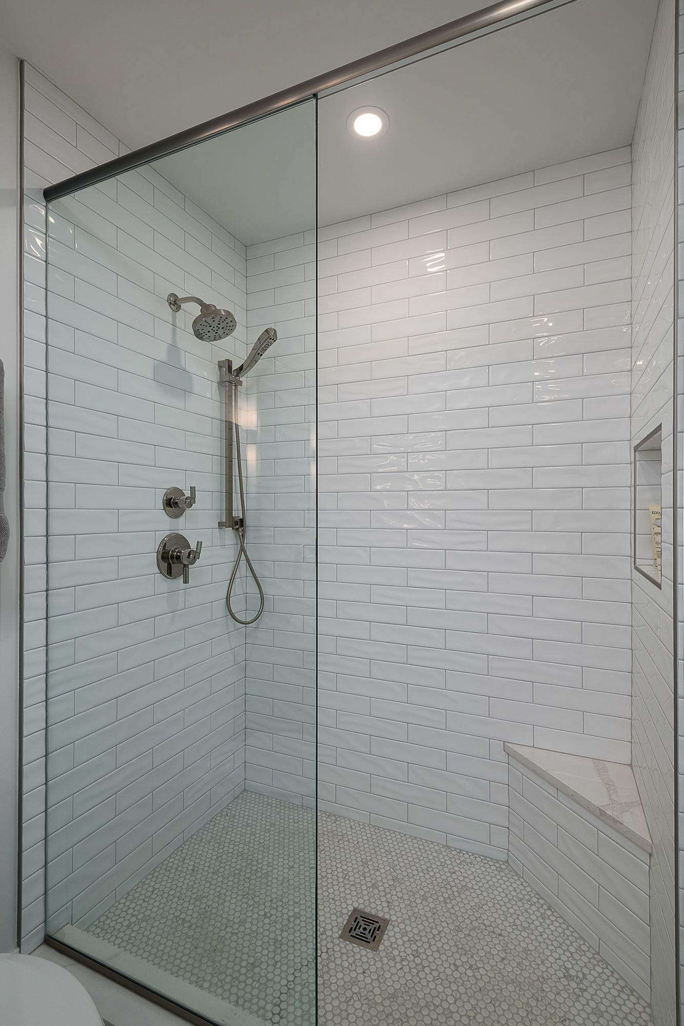 A bathroom with white tiles and a walk in shower with a glass door.