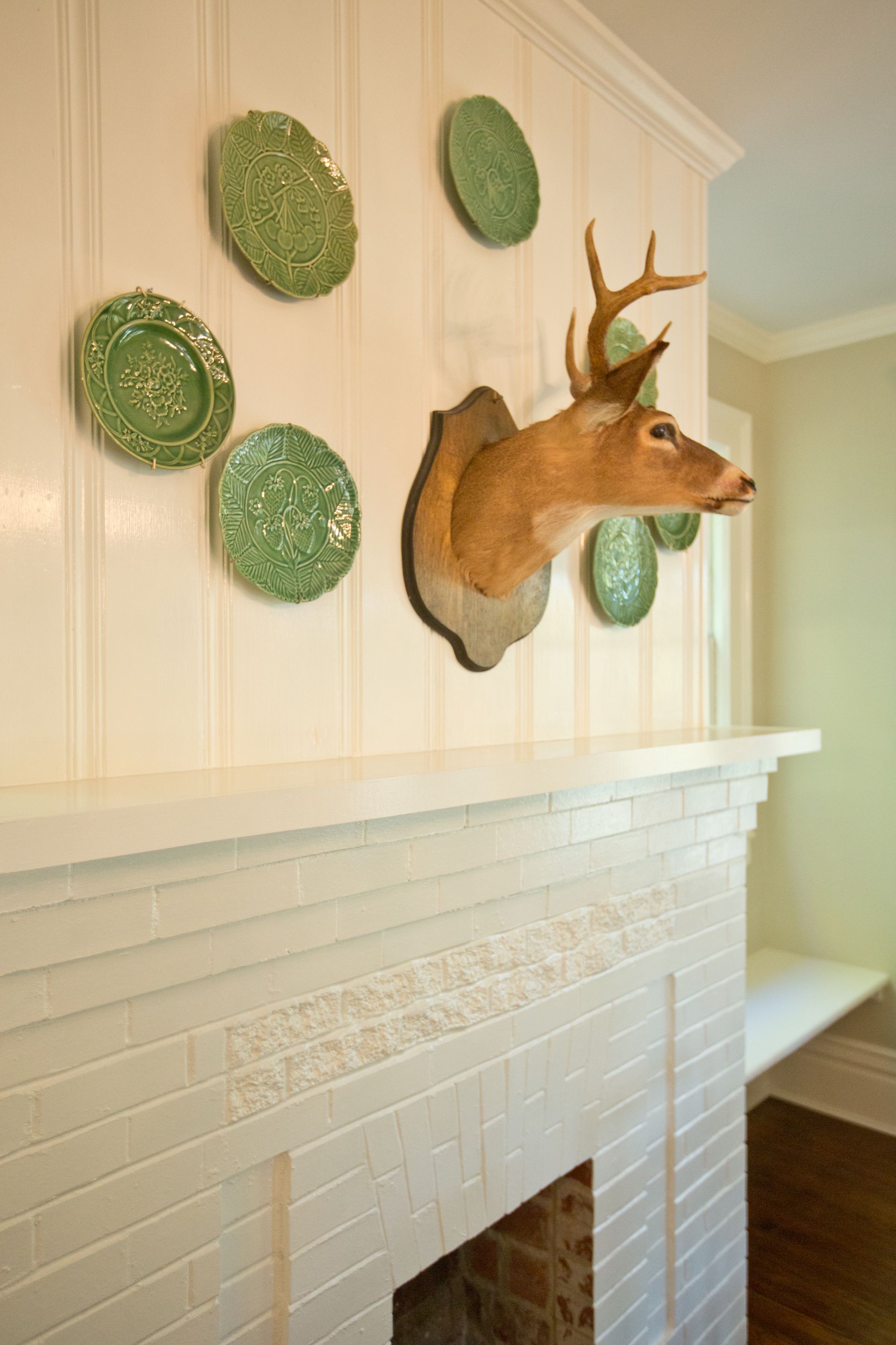 A deer head is hanging on a wall above a fireplace surrounded by green plates.
