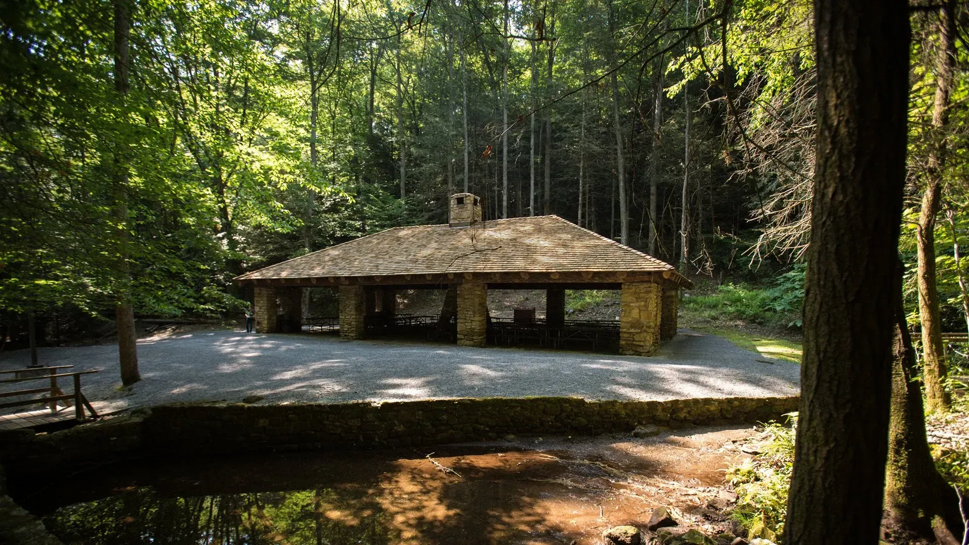A stone pavilion in the middle of a forest with a pond in front of it.