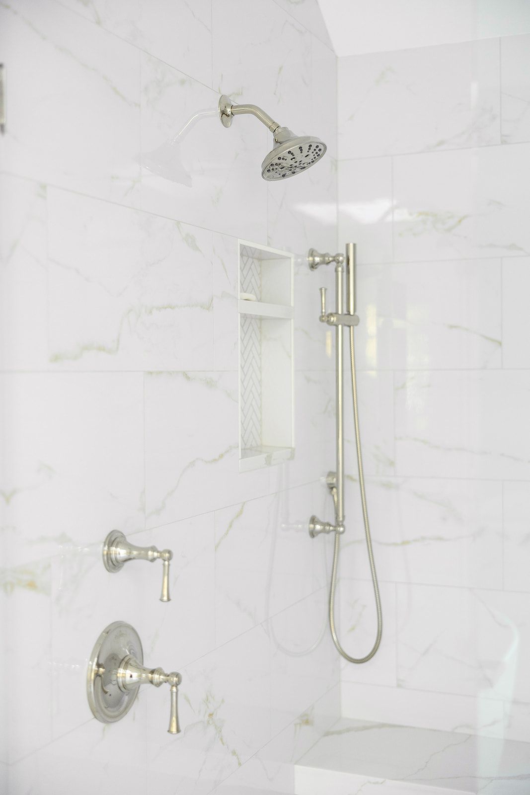 A shower with a stainless steel shower head and faucets on a white tiled wall.