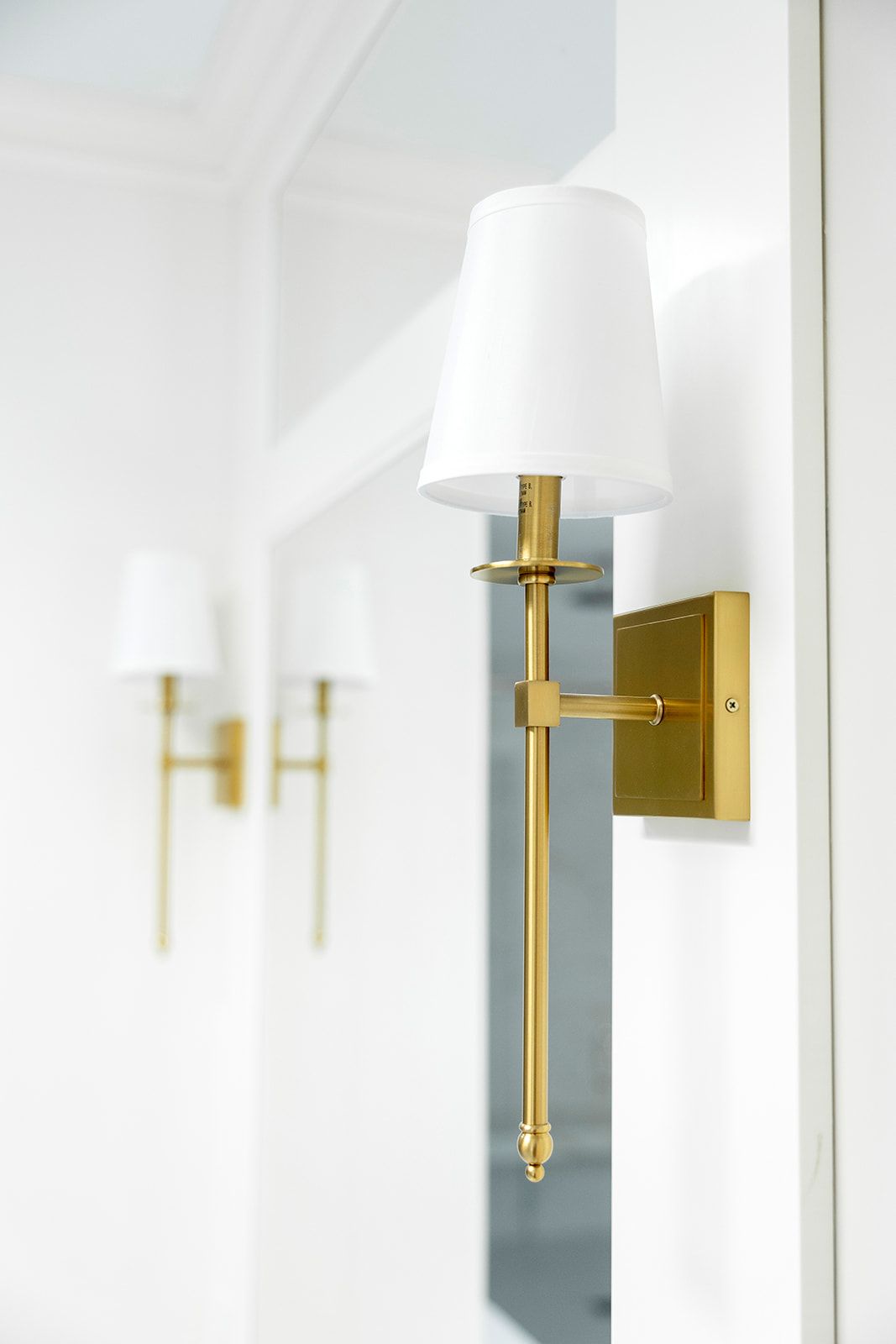 A brass wall light with a white shade is hanging on a white wall.