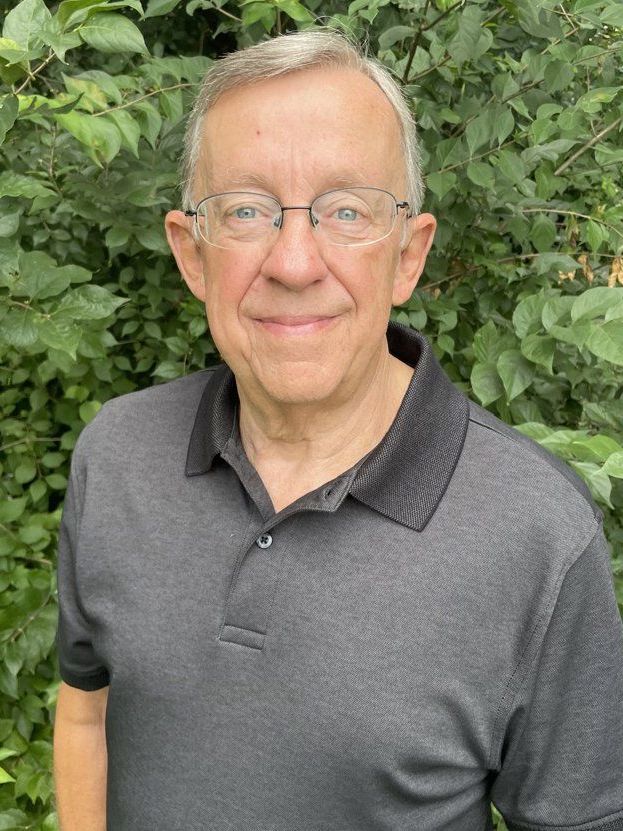 A man wearing glasses and a black shirt is standing in front of a bush.