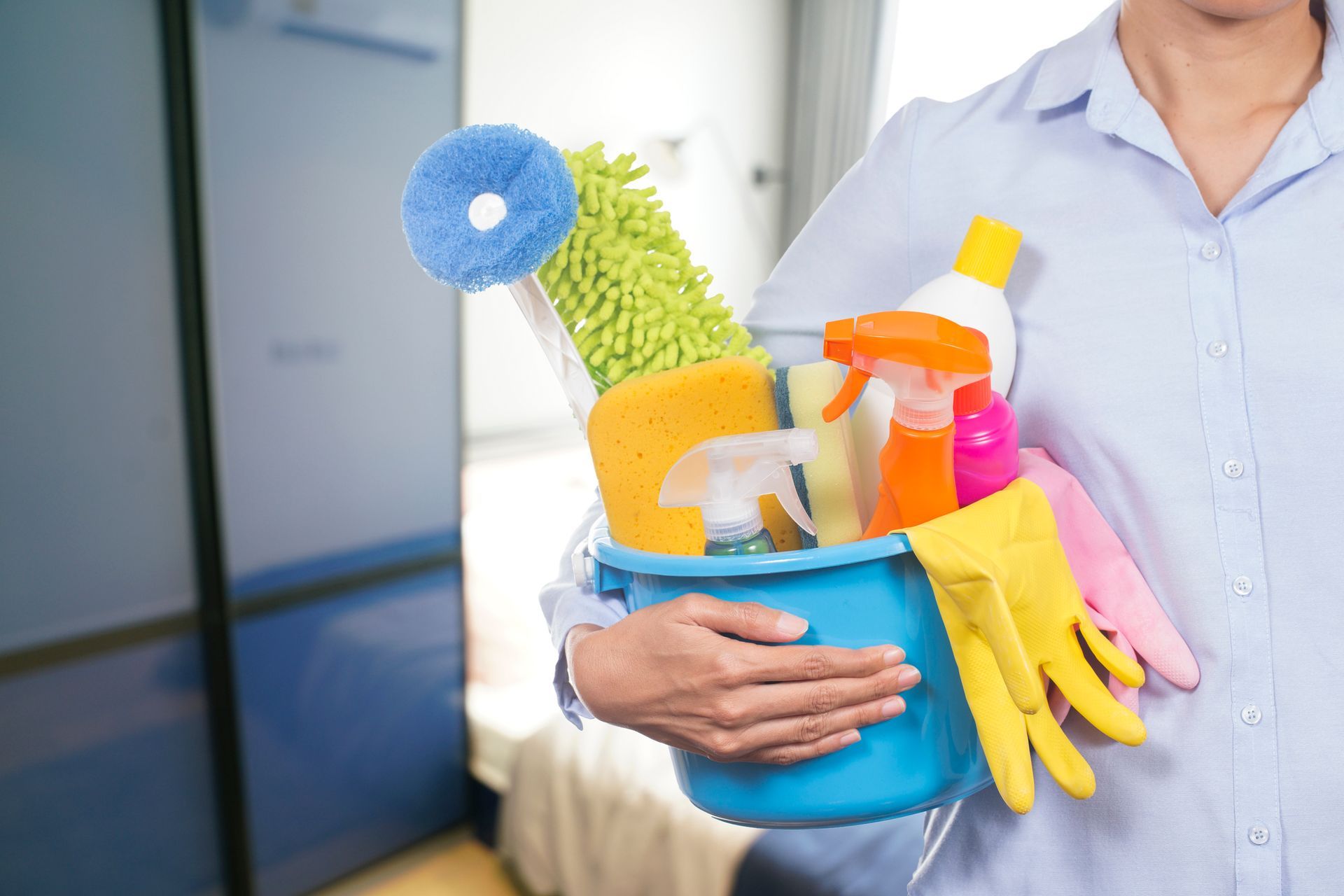 Person holding a blue bucket filled with cleaning supplies, including a mop, sponges, and spray bottles.