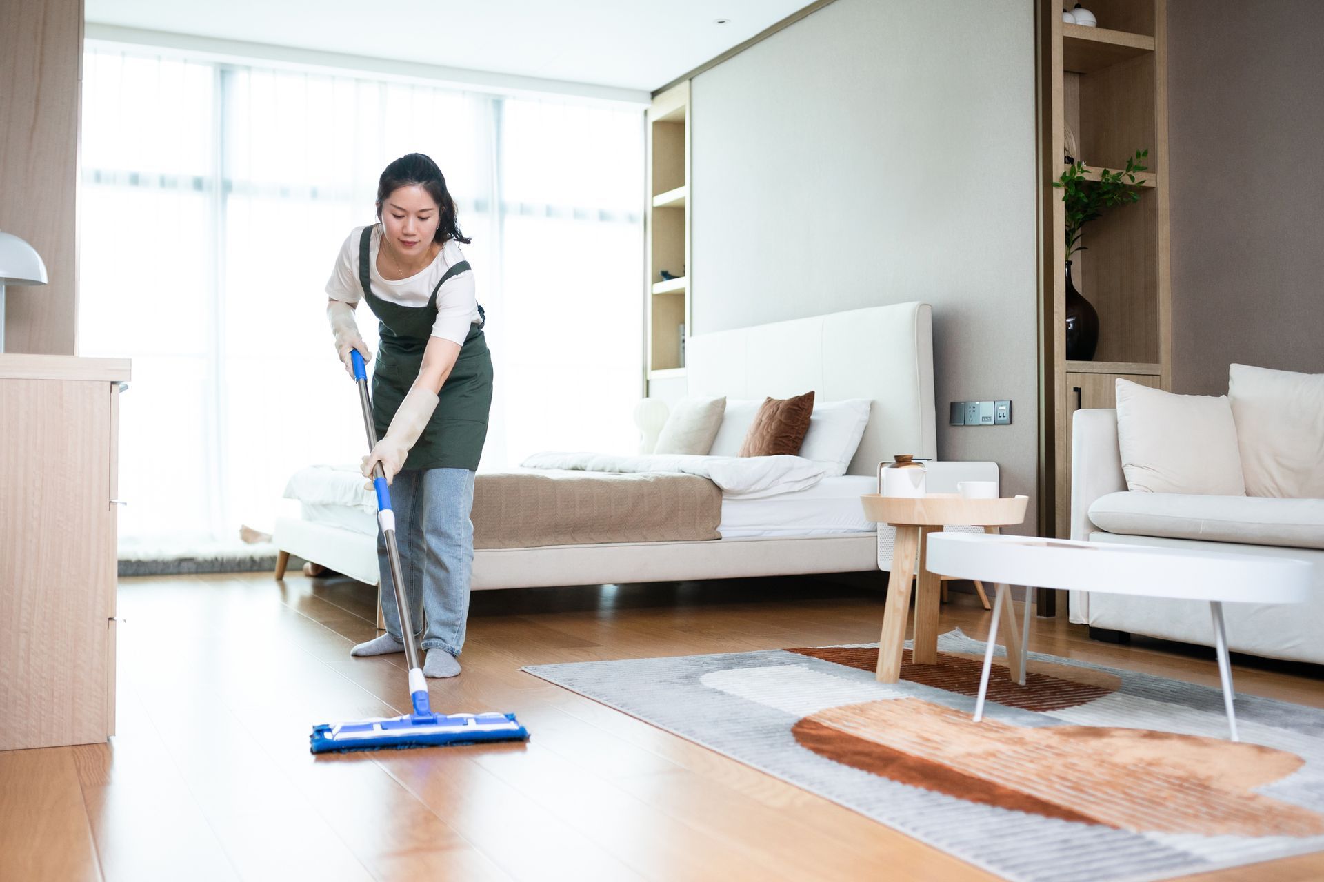 Woman mopping a wooden floor in a bedroom with a bed, sofa, and rug.