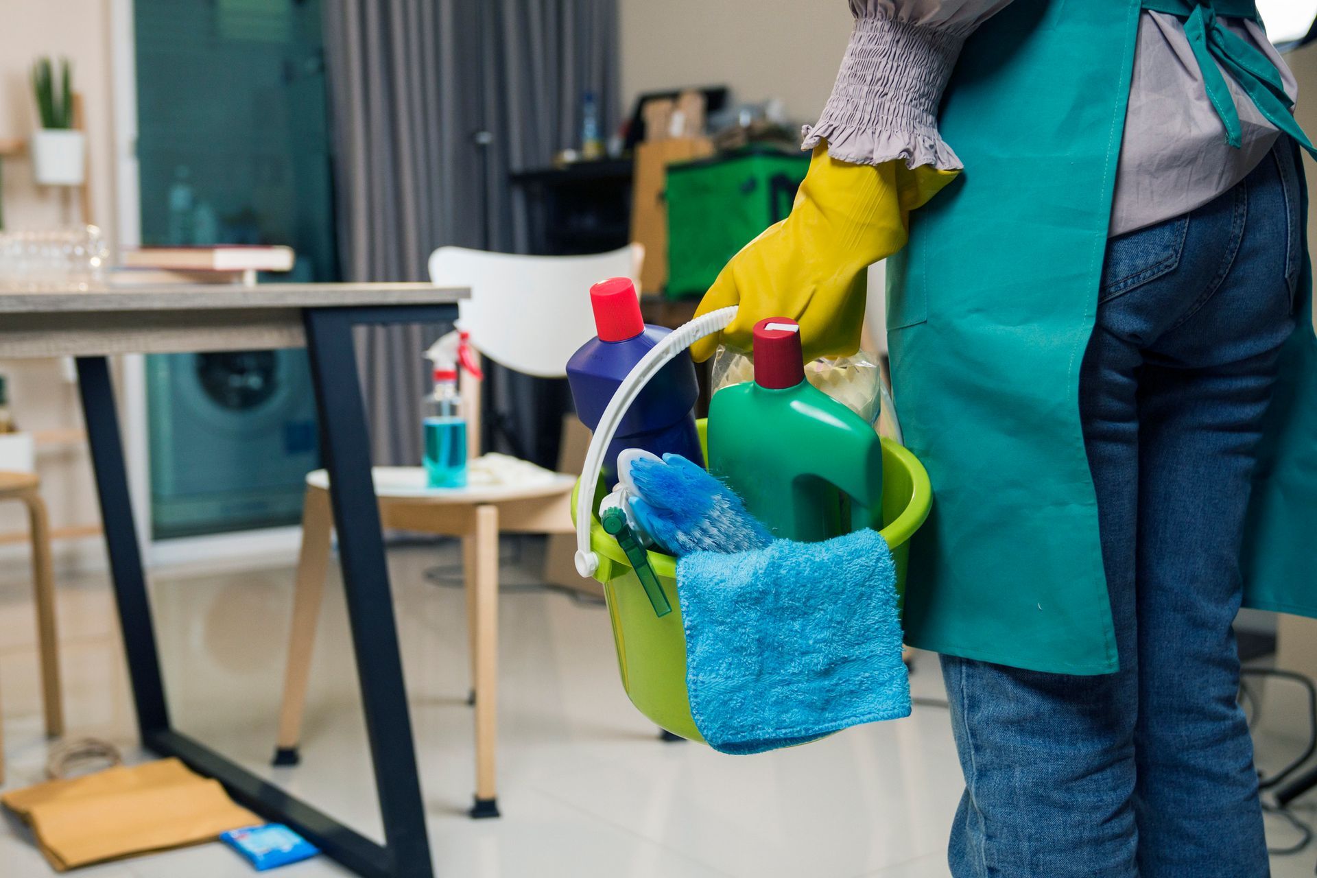 Person holding cleaning supplies in a bucket, standing in a messy room.