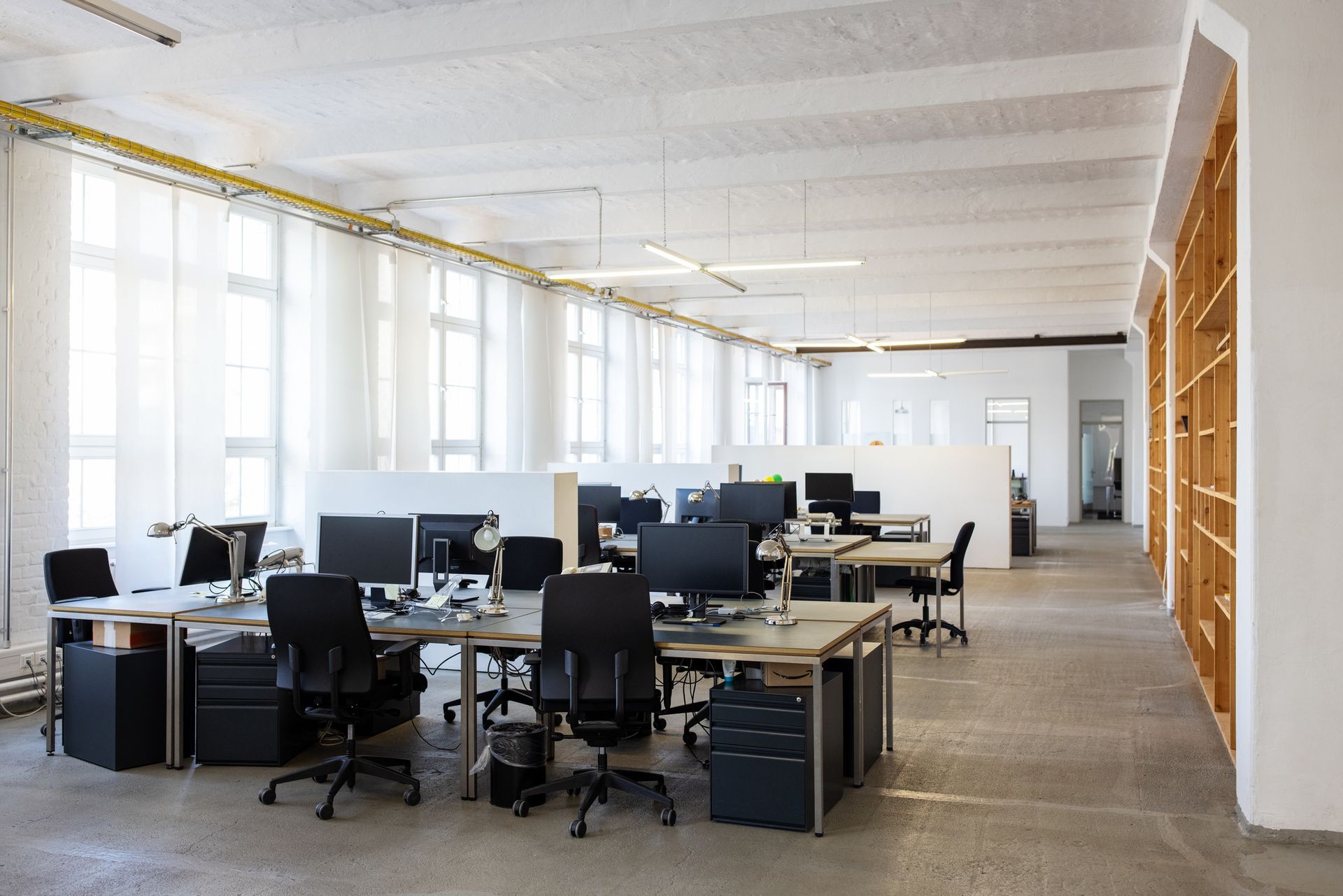 Open-plan office with multiple computer workstations, tall windows with white curtains, and a concrete floor.