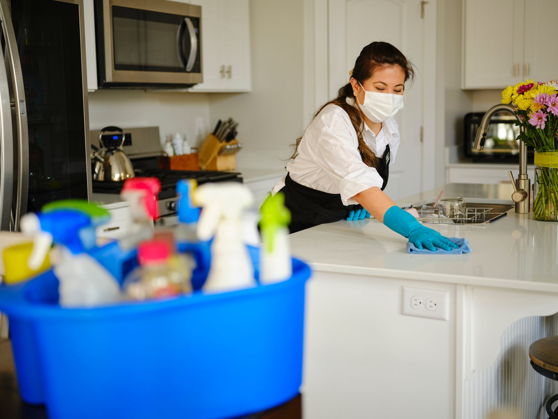 Person wearing mask and gloves cleaning a white countertop in a kitchen; cleaning supplies in blue bucket nearby.