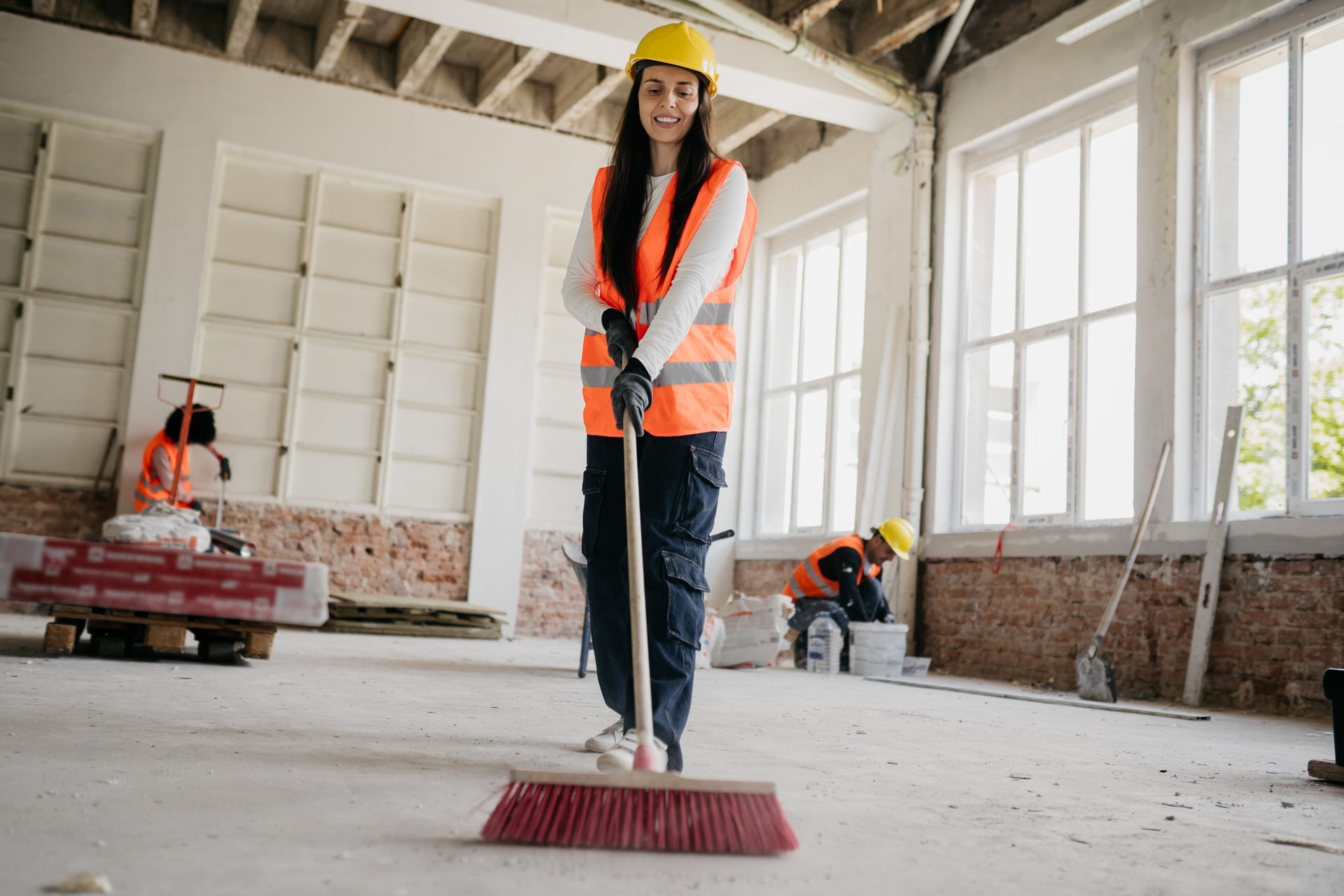Woman sweeping a construction site, wearing safety gear and smiling. Two workers are in the background.