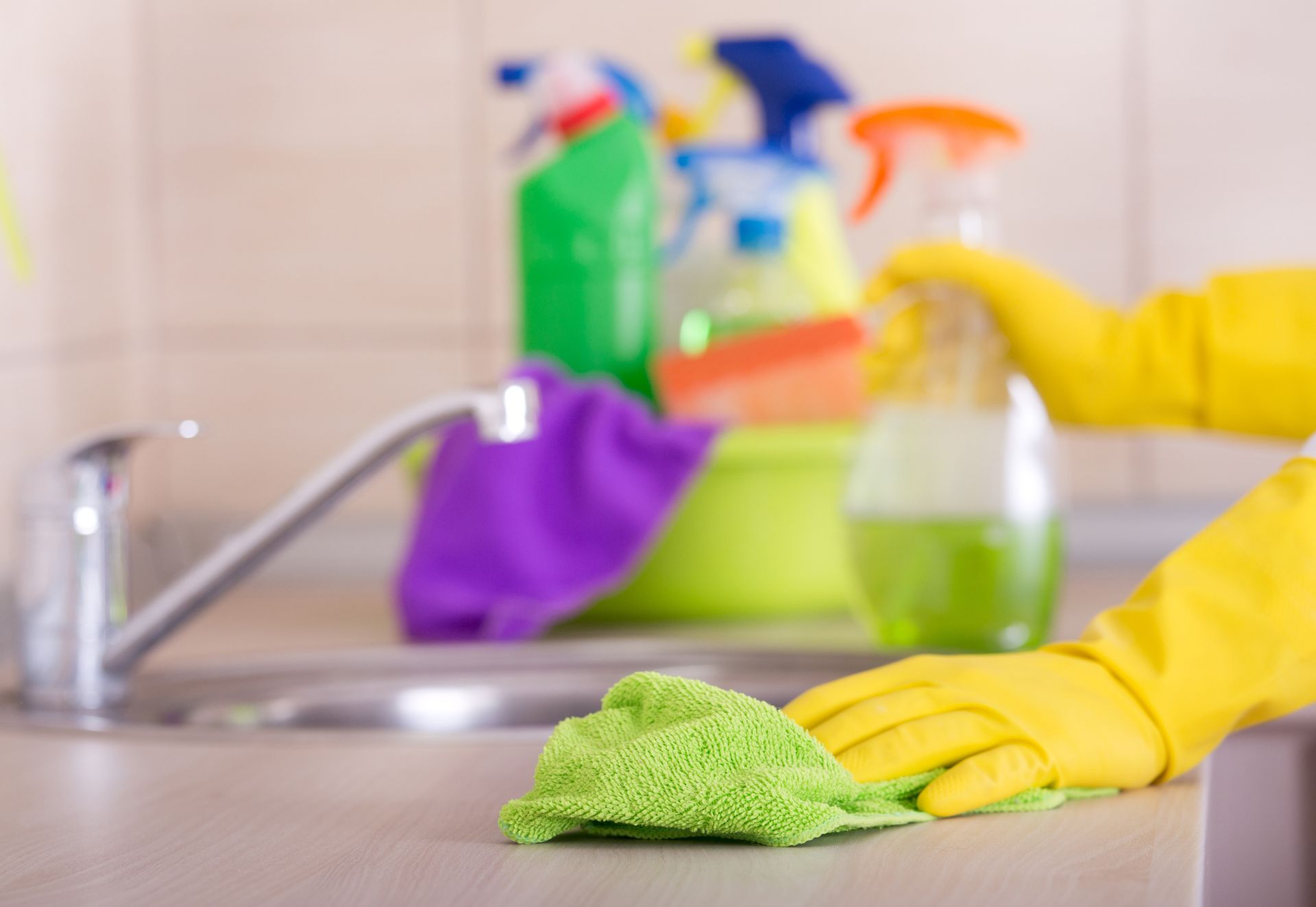 Person wearing yellow gloves wiping a kitchen countertop with a green cloth, cleaning supplies in the background.