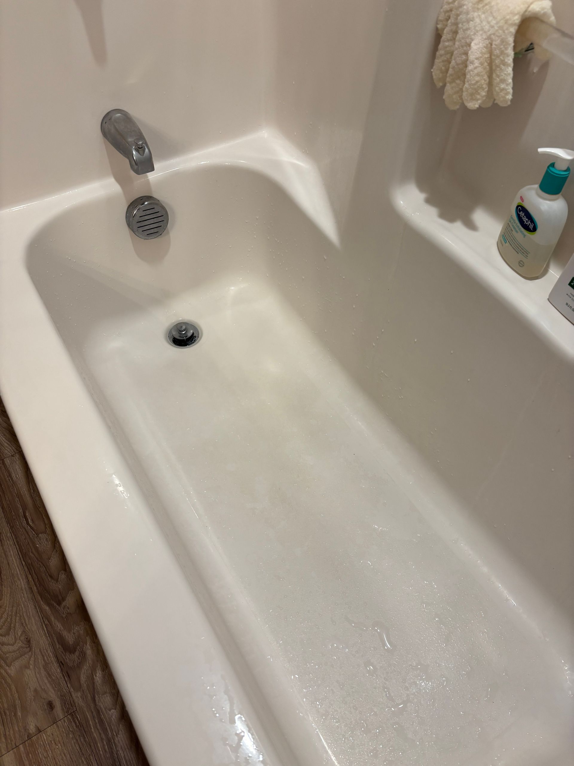 Two people in blue cleaning uniforms in a kitchen, one smiling with arms crossed, holding cleaning supplies.