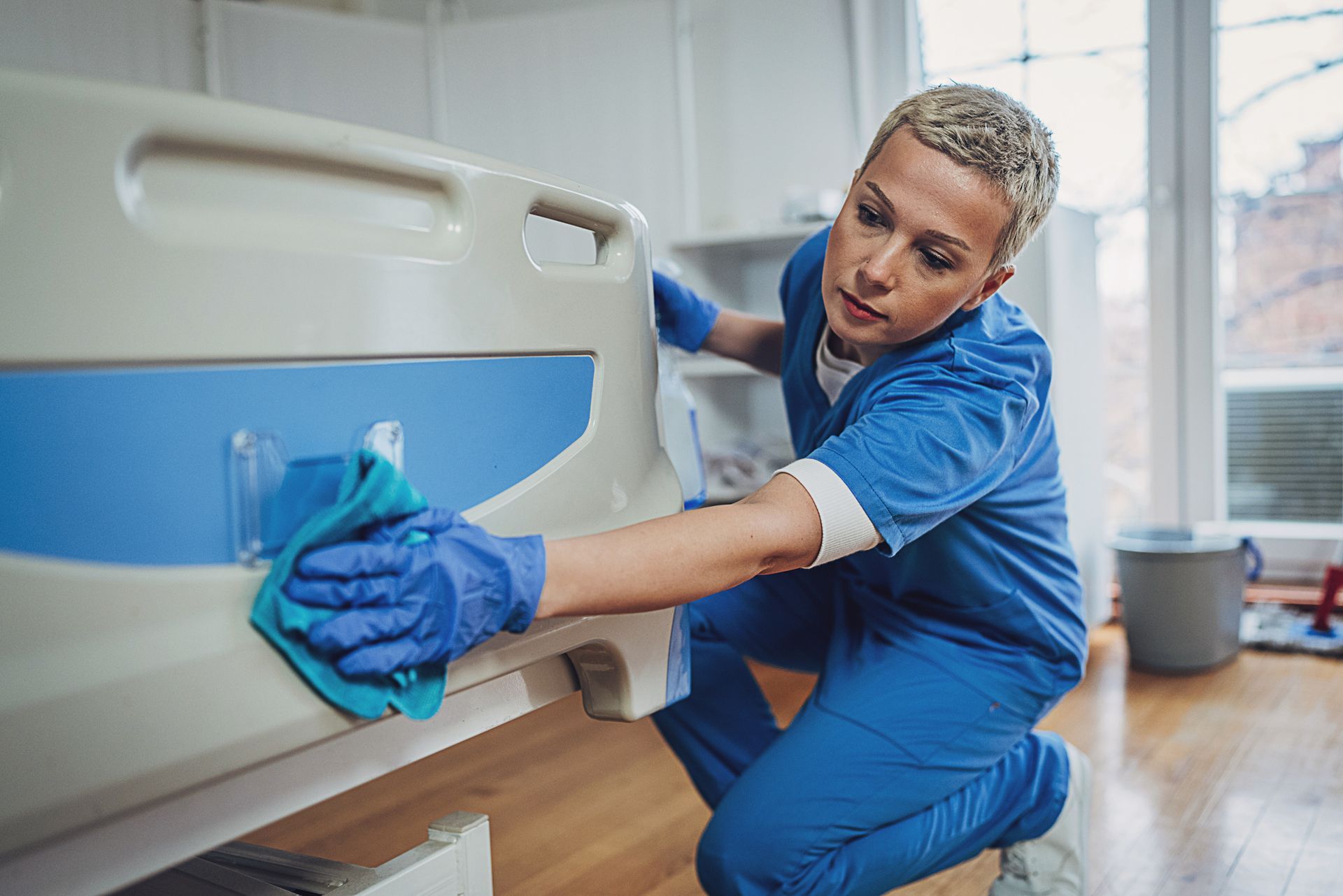 Person in blue scrubs cleans a hospital bed with a blue cloth.