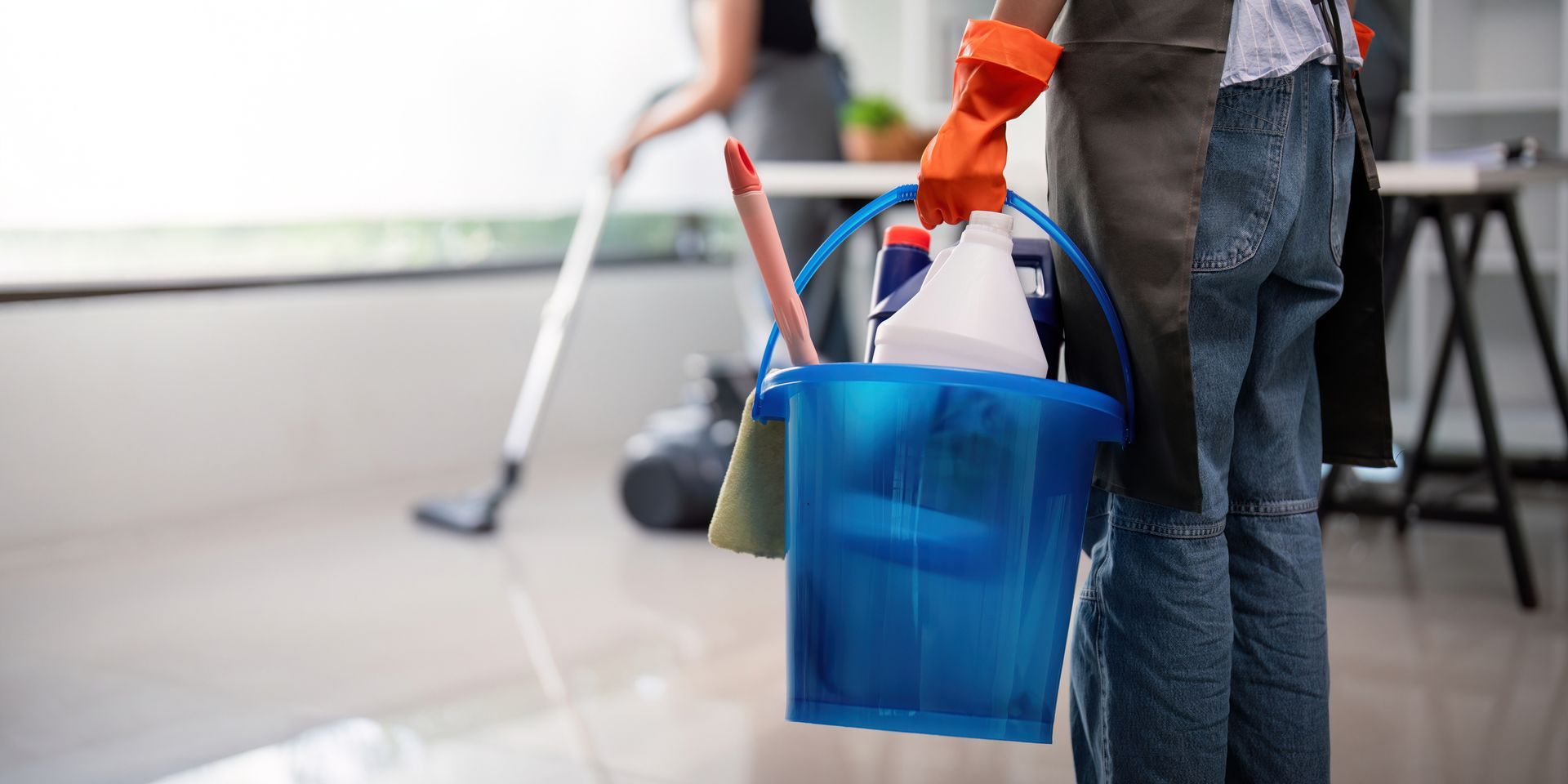 Person holding a blue cleaning bucket with gloves on, vacuuming a room.