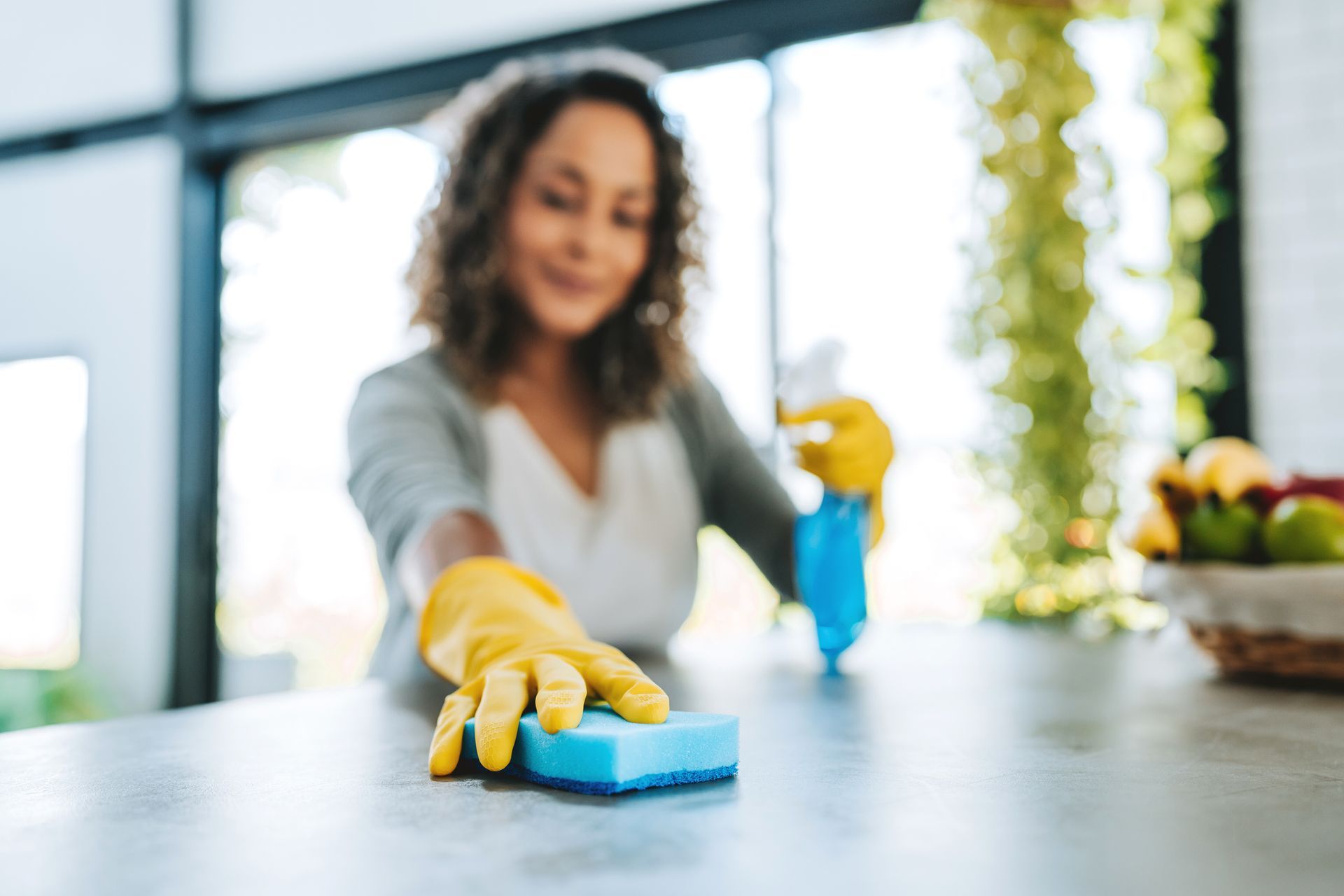 Woman wearing gloves cleans kitchen counter with a sponge and spray bottle.