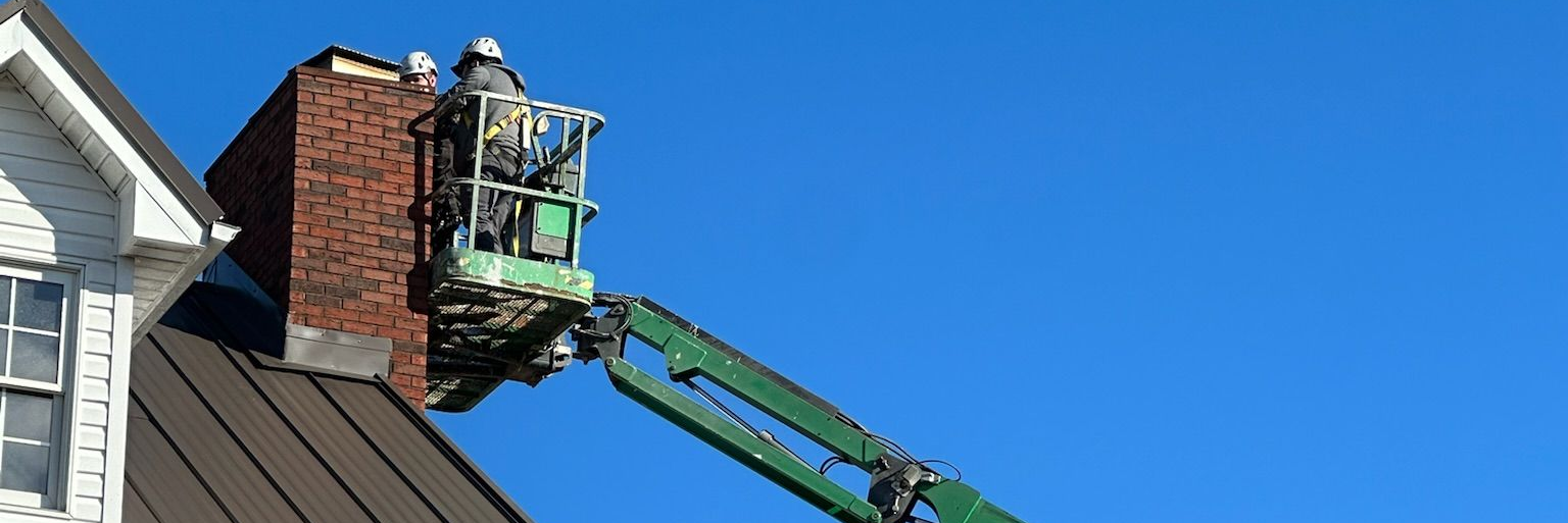 Certified fireplace technician on a green boom lift repairing a chimney on a house roof against a blue sky