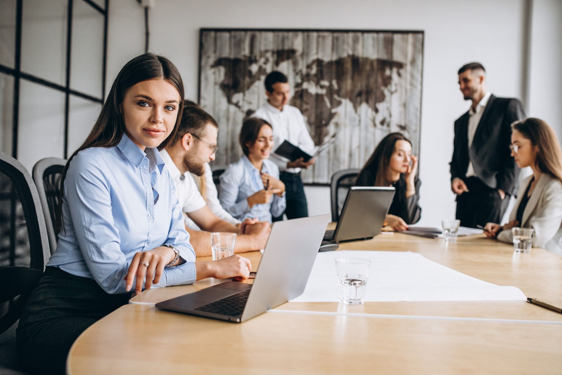 Reunião de negócios: Mulher à cabeceira de uma mesa com colegas, olhando para a câmera. Laptop e mapa-múndi ao fundo.
