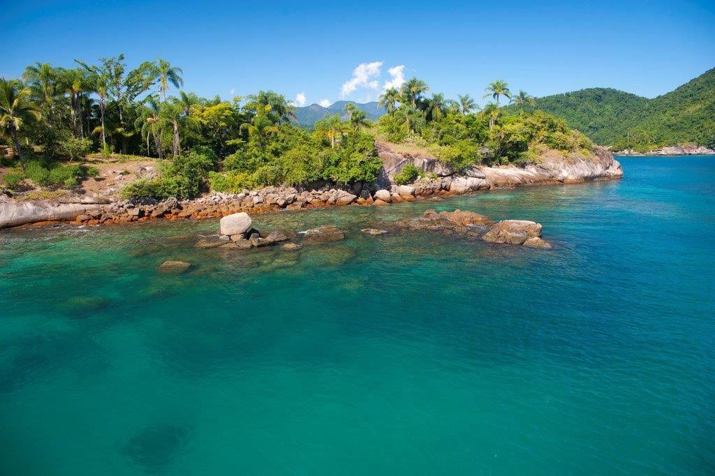 Água esmeralda e litoral rochoso, árvores verdejantes, céu azul. Cenário costeiro tropical.