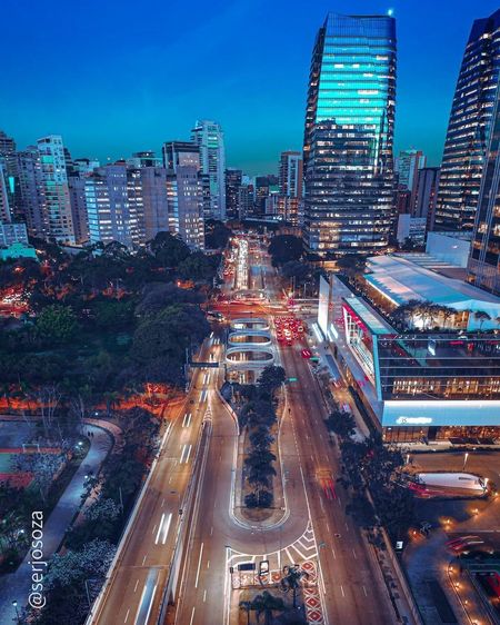Rua da cidade ao anoitecer, prédios altos iluminados, estradas movimentadas com trilhas iluminadas, estacione à esquerda.