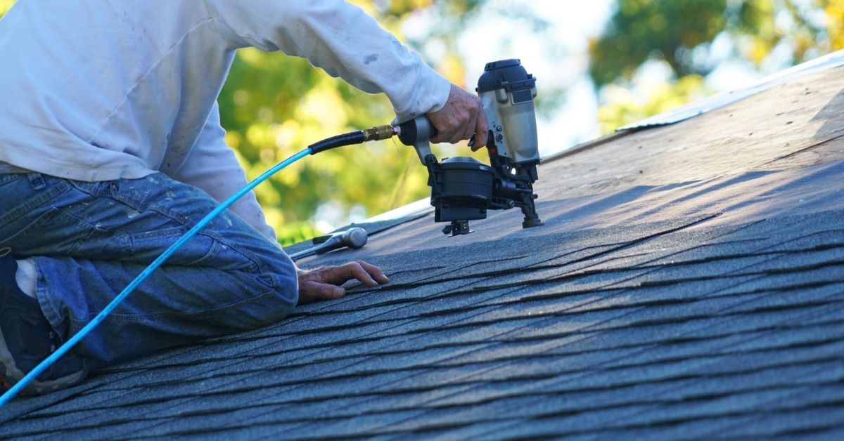 A roofer in jeans and a white shirt kneels on a residential roof, using a nail gun to install asphalt shingles.