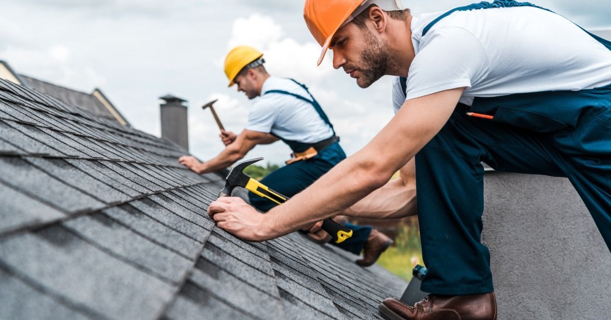 Two roofers in safety gear install shingles on a sloped residential roof, using hammers under a cloudy sky.