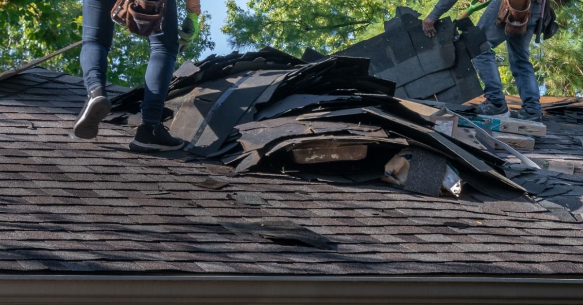 Two roofers remove damaged shingles during a roof tear-off, with debris piled high on a residential 
