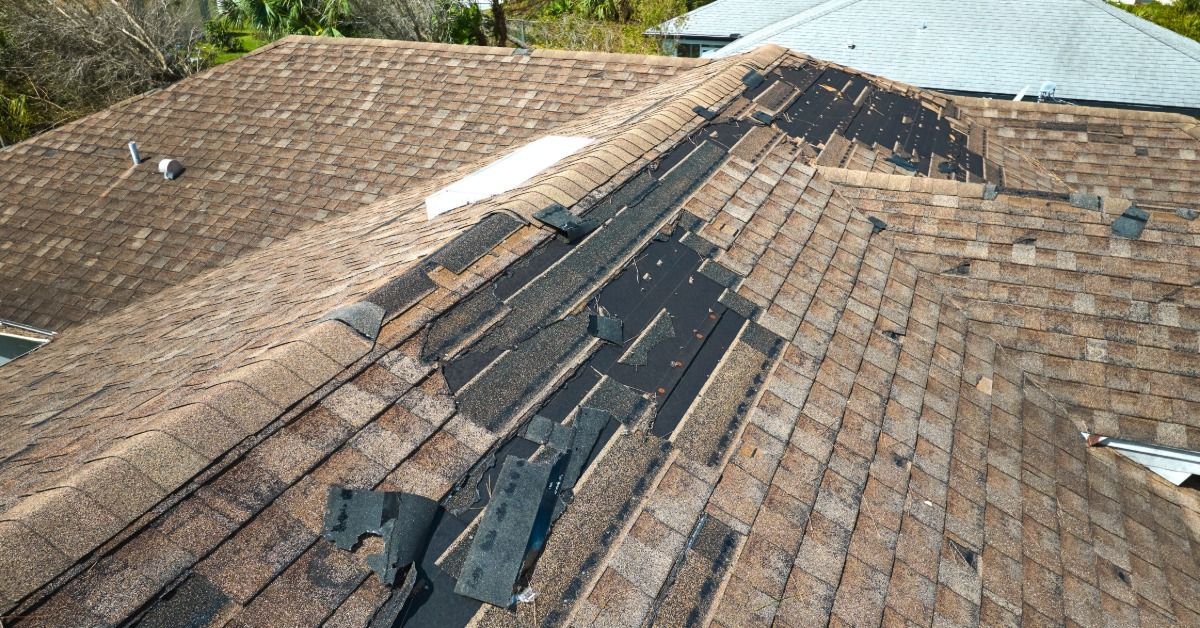 A wind-damaged asphalt roof with torn shingles and exposed underlayment across multiple roof section