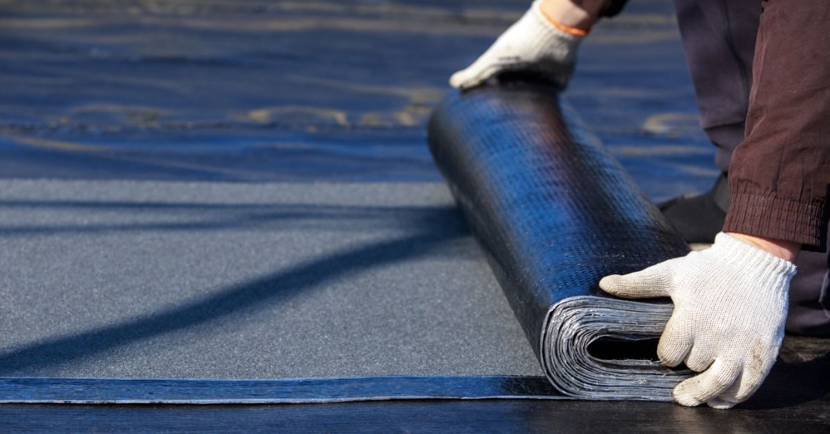 A worker wearing gloves rolls out a black EPDM roofing membrane across a flat commercial roof during