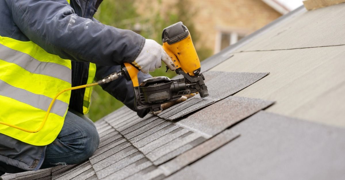 A roofing contractor installs gray asphalt shingles with a nail gun while working on a sloped reside