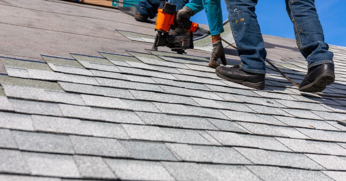 Roofing contractors install gray asphalt shingles on a sloped roof, using a nail gun during a roof replacement project.